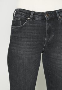 Gros plan sur un jean en denim noir porté avec un haut blanc côtelé rentré, montrant les poches avant, les passants de ceinture et une fermeture bouton métal.