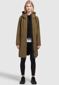 Manteau imperméable vert olive avec capuche, fermeture éclair et poches latérales. Porté par-dessus un haut noir et des leggings, avec un design élégant et moderne.