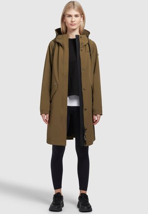 Olive green waterproof coat with a hood, zip closure, and side pockets. Worn over a black top and leggings, featuring a sleek, modern design.