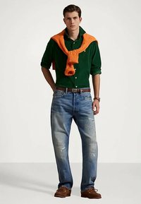 Zielona koszula zapinana na guziki, pomarańczowy sweter zarzucony na ramionach, wyblakłe niebieskie dżinsy z przetarciami, brązowe buty i brązowy pasek.