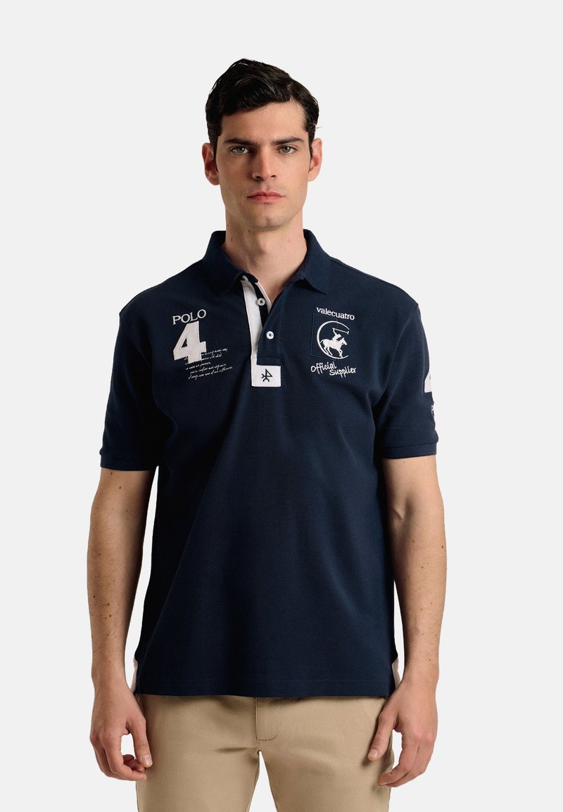 Camisa polo marina de manga corta, con cuello blanco, logo bordado y detalle del número 4. Presenta una tela texturizada y un ajuste entallado.