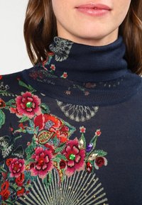 Marineblauer Rollkragenpullover mit bunter Blumenstickerei, Schmucksteinakzenten und einem strukturierten Strickdesign.