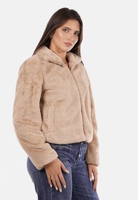 Giacca in pelliccia finta beige con zip e colletto, caratterizzata da una finitura morbida e testurizzata e tasche laterali, indossata con jeans blu.