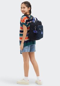 Sac à dos en nylon bleu avec un logo rond, poche zippée à l'avant et porte-clés en pom-pom décoratif. Modèle portant un pull rayé et un short en denim.
