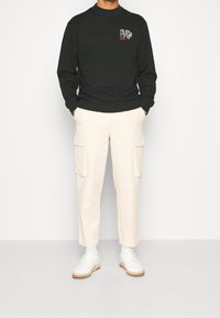 Felpa nera con collo a giro e logo grafico sul petto, abbinata a pantaloni cargo beige con tasche laterali e sneakers bianche.