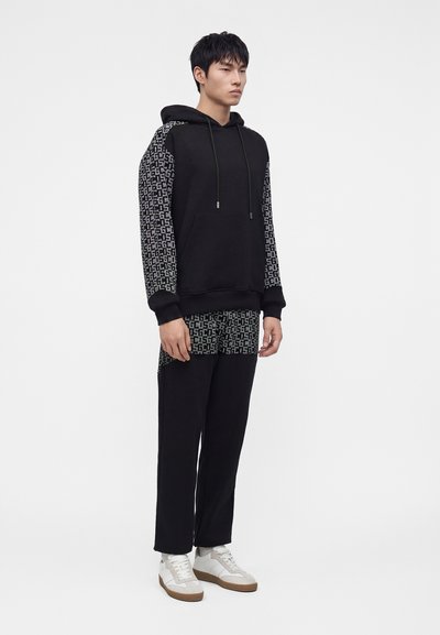 Homme en sweat à capuche noir avec manches à motifs et pantalon assorti, debout, portant des baskets blanches sur un fond blanc uni.