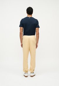 Άνδρας π ου στέκεται με την πλάτη προς τα πίσω, φοράει μπλε σκούρο μπλουζάκι, μπεζ φόρμες και λευκά αθλητικά παπούτσια μπροστά σε ένα απλό λευκό φόντο.