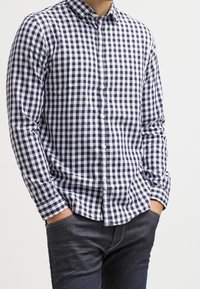 Camicia a quadri blu navy e bianca con maniche lunghe, colletto e pattina frontale. Indossata con jeans scuri, che evidenziano un fit rilassato.