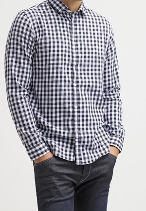 Camisa de cuadros en azul marino y blanco, de botones, con mangas largas, cuello y tapeta frontal. Se lleva con jeans oscuros, mostrando un ajuste relajado.