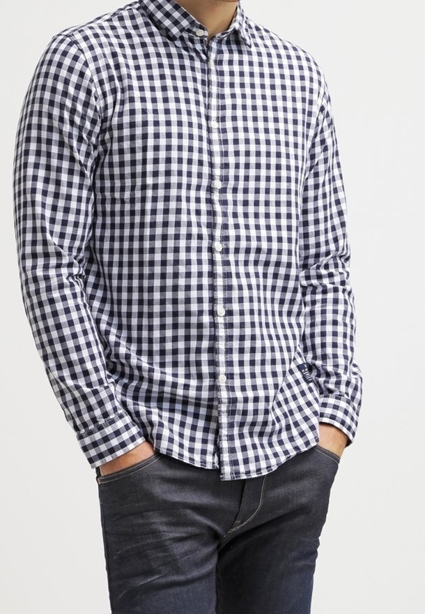 Camicia a quadri blu navy e bianca con maniche lunghe, colletto e pattina frontale. Indossata con jeans scuri, che evidenziano un fit rilassato.