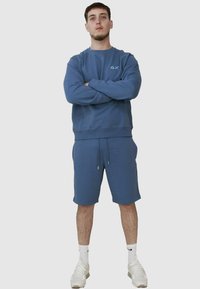 Giovane uomo in piedi con le braccia incrociate, indossa una felpa blu e pantaloni corti coordinati, calzettoni bianchi e sneakers bianche, su uno sfondo uniforme chiaro.