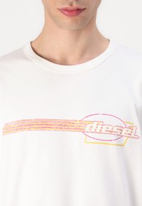 White cotton T-shirt featuring a pink and yellow graphic design with "diesel" text, round outline, and horizontal striped accents.