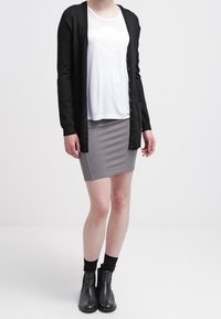 Cardigan nero sopra una camicia bianca con dettagli in pizzo, abbinato a una gonna a matita grigia, stivaletti neri e calzini scuri.