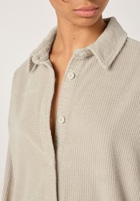 Gaiši pelēka kordurojas krekls ar klasisku apkakli, ar pogu aizdari un vertikālu ribu faktūru. Auduma detaļu tuvplāns.