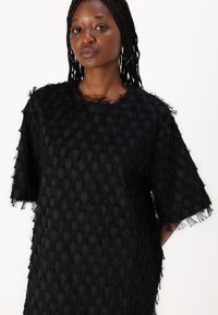 Haut noir à manches courtes, texturé avec un motif à franges, avec un col rond et une coupe ample. Le tissu semble doux et superposé.