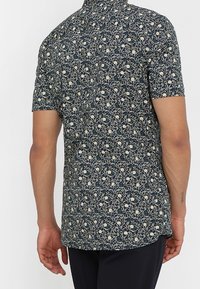 Camicia a maniche corte in blu navy scuro con un motivo floreale che presenta fiori color crema e foglie verdi; tessuto traspirante con una vestibilità comoda.