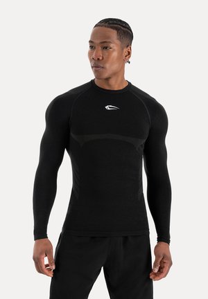 Atletyczny mężczyzna z zaplecionymi włosami, ubrany w dopasowaną czarną bluzę z długim rękawem i czarne spodnie, stojący i patrzący w bok na białym tle.