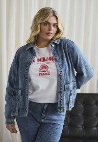 Jeansjacka med två framfickor, knappstängning och långa ärmar, bärs över en vit t-shirt med röd text. Blå jeans fulländar outfiten.