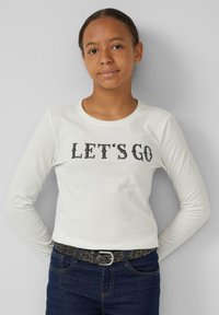 Haut blanc à manches longues avec "LET'S GO" imprimé en noir. Accompagné d'un jean en denim foncé et d'une ceinture texturée noire et grise.