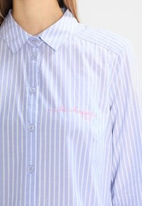 Helles blau gestreiftes Hemd mit Knöpfen und Kragen, mit rosa gesticktem Text "be happy" im linken Brustbereich. Leichter Stoff.