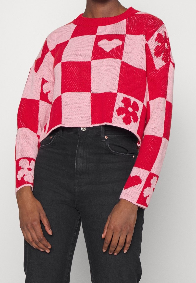 Femme portant un pull court à carreaux rouge et rose avec des motifs de cœurs et de fleurs, associé à un jean noir taille haute.
