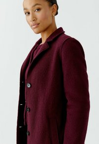 Burgundy wool coat with textured fabric, notched collar, and black buttons. Features side pockets and a tailored fit.