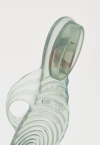 Gjennomsiktig, fleksibel plastkanne med buede ribber, ergonomisk håndtak og rund tut. Inkluderer preget merkenavn på bunnen.