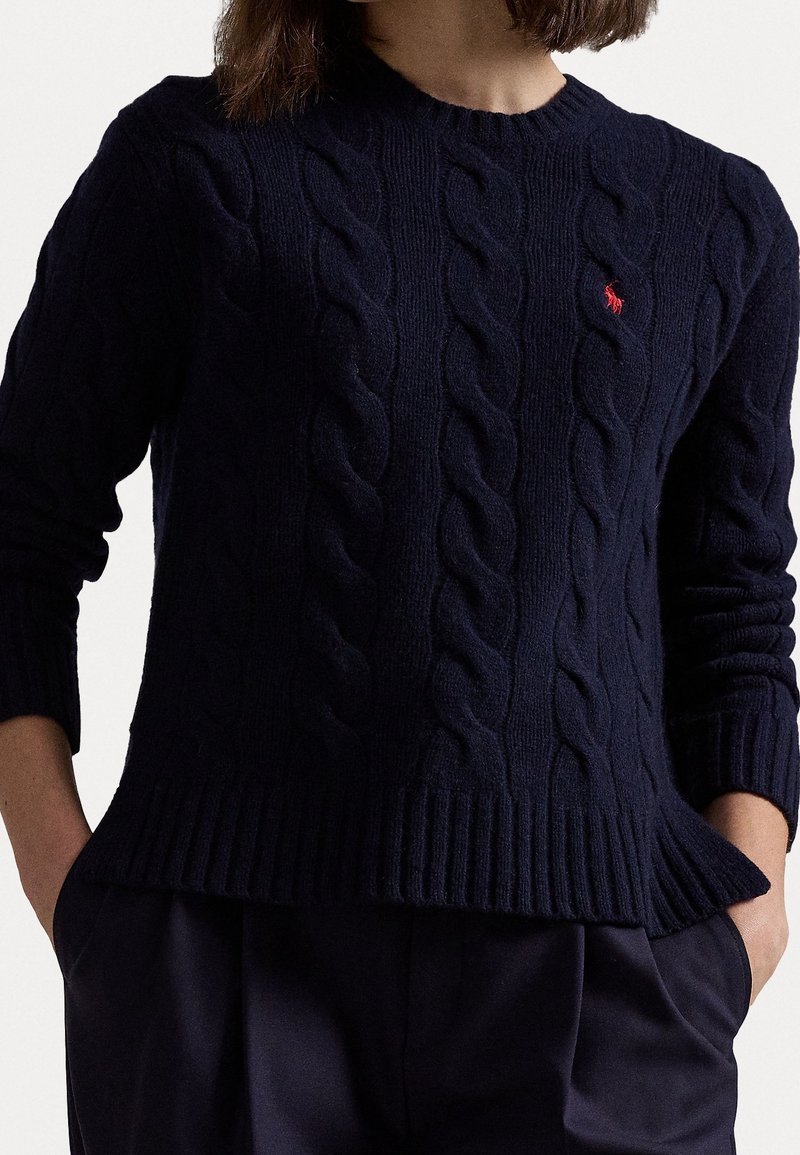 Mørkeblå strikket sweater med kabelmønster, ribbet halsudskæring og kant. Har et lille rødt broderet logo på brystet. Lange ærmer.