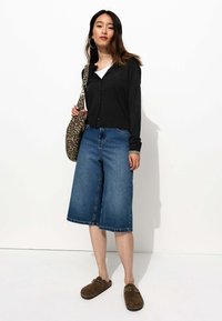 Gilet noir à boutons sur un T-shirt blanc, associé à des culottes en denim bleu à jambes larges et des sabots marron. Sac à épaule en imprimé léopard.