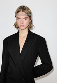 Schwarzer Blazer mit tiefem V-Ausschnitt, ausgestattet mit einer glatten Textur und strukturierten Schultern. Akzentuiert durch ein silbernes Ketten-Haarteil.