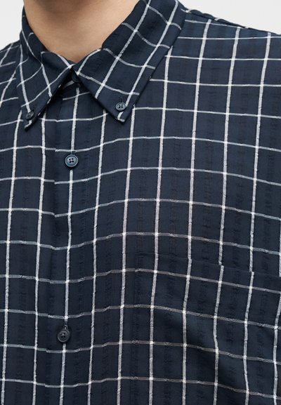 Πουκάμισο σε ναυτικό μπλε χρώμα με λευκό πλέγμα, διαθέτει γιακά με κουμπιά και τσέπη στο στήθος.