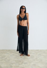 Črn bikini zgornji del s detajlom vozliča, povezan z zračnimi črnimi nabranimi hlačami. Očala za sonce nošena, bosih nog na teksturiranem kamnitrem tleh.