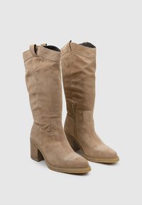 Botas de ante beige hasta la rodilla con punta redondeada, detalles de costura y cremallera lateral, que cuentan con un tacón cuadrado y acentos decorativos.