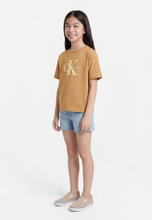 Pige stående og smilende, iført en brun Calvin Klein t-shirt, lyse denimshorts og beige slip-on sko foran en hvid baggrund.