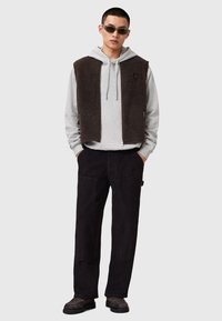 Outfit da uomo con una felpa grigia con cappuccio e lacci, un gilet in pile marrone scuro e pantaloni in velluto a coste neri. Indossato con occhiali da sole e scarpe casual.