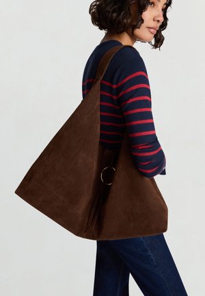 Braune Wildleder-Schultertasche mit dreieckiger Form, großem goldfarbenem Ringakzent und weicher Textur. Das Modell trägt ein gestreiftes Oberteil in Marineblau und Rot.