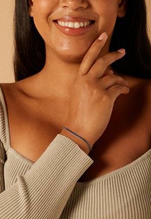 Bracelet bleu en perles sur un poignet bronzé, avec de petites perles rondes sur une chaîne délicate. Le décor est minimaliste avec des tons neutres.