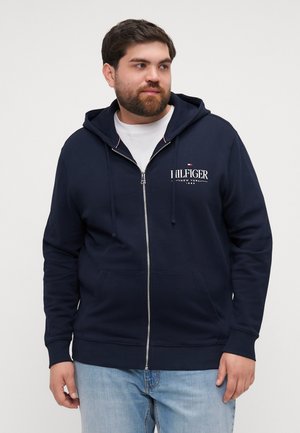 Mornarica zip-up dukserica napravljena od mekanog materijala, sa kenguru džepom, vezicama i detaljima loga sprijeda u bijeloj boji.