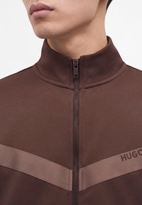 Gros plan d’une personne portant une veste marron zippée avec un col montant et une bande horizontale marron plus claire sur la poitrine.