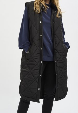 Personne portant un long gilet matelassé noir sur un sweat bleu marine et un pantalon noir, les mains dans les poches du gilet.