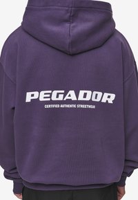 Ljubičasta dukserica s velikim bijelim logotipom "PEGADOR" i natpisom "CERTIFIED AUTHENTIC STREETWEAR" na leđima. Mekani, teksturirani materijal.