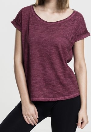 Woman wearing a short-sleeve, rolled-cuff, heathered burgundy T-shirt and black pants, posing with one hand resting on thigh.