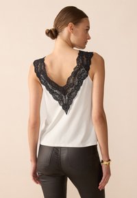 Top blanco sin mangas con un escote en V profundo, que presenta un borde de encaje negro a lo largo del cuello. Combinado con pantalones de cuero negros.