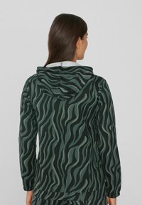 Una mujer con el pelo largo y marrón lleva una chaqueta con capucha a rayas de cebra en negro y verde oscuro, vista desde atrás contra un fondo gris liso.