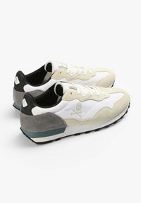 Sneaker mit einem weißen Mesh-Obermaterial und cremefarbenen Wildlederakzenten, ausgestattet mit einer schwarzen und grauen Ferse, hexagonalem Logo und schwarzem Gummisohle.