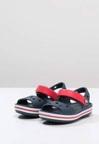 Sandales bleu marine avec des brides ajustables rouges, présentant des trous de ventilation et des semelles rayées rouges et blanches pour une bonne adhérence et un confort optimal.