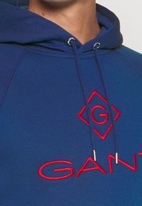 Sudadera azul marino con un bolsillo frontal, que presenta un logotipo y texto bordados en rojo. Los cordones tienen puntas metálicas de color plata. Tejido suave y texturado.
