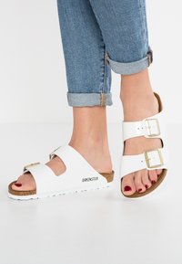 Sandalias Birkenstock blancas con dos correas ajustables, plantilla de corcho y una suela blanca texturizada, combinadas con unos vaqueros azules remangados.