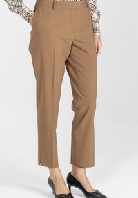 Kobieta w camelowych, dopasowanych spodniach, w koszuli w kratę i czarnych szpilkach, stojąca z jedną ręką w kieszeni na białym tle.