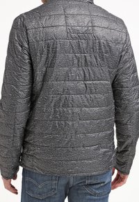 Grå quilted puffer jakke med en tekstureret overflade, der har vandrette sømme og en opretstående krave. Båret med blå jeans.